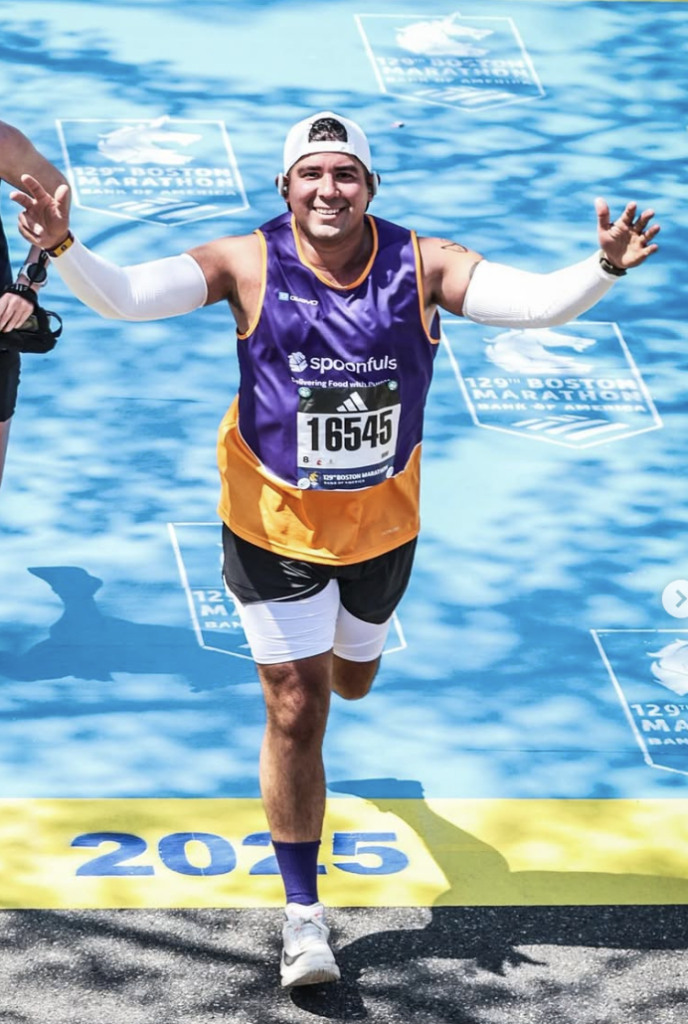 A member of Spoonfuls' 2025 Boston Marathon team crossing the finish line smiling with his arms raised high.